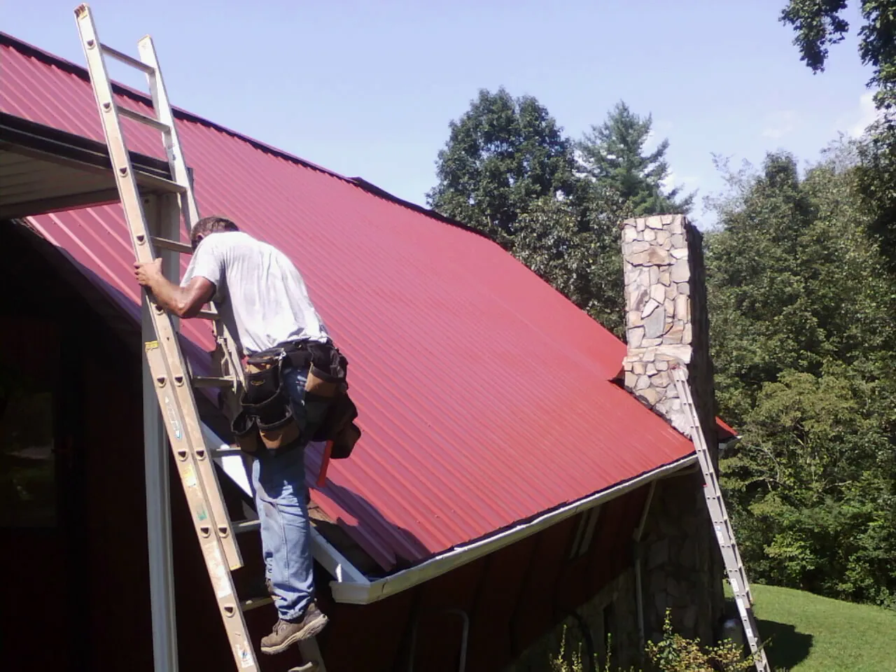 Roofer on a ladder beside a red metal roof and stone chimney — Soffit & Fascia Repair by Roofing Brothers in Hazel Green